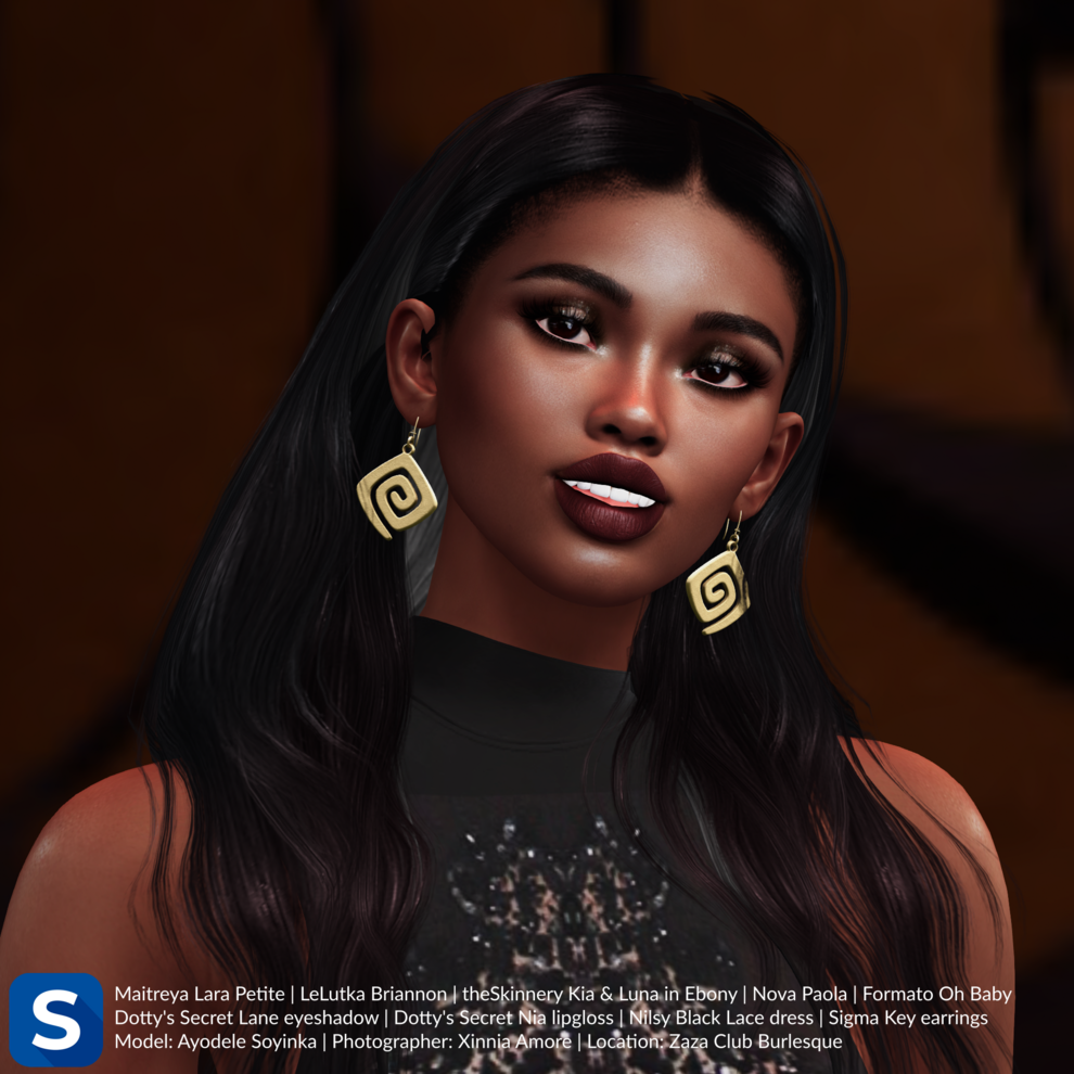 A headshot of Ayodele Soyinka standing in front of a wall with brown-and-black patterned wallpaper. She is wearing a black-lace sleeveless dress and gold pendant earrings, while her black hair is long and straight. Her eyeshadow is brown and smokey while her lip gloss is dark red.