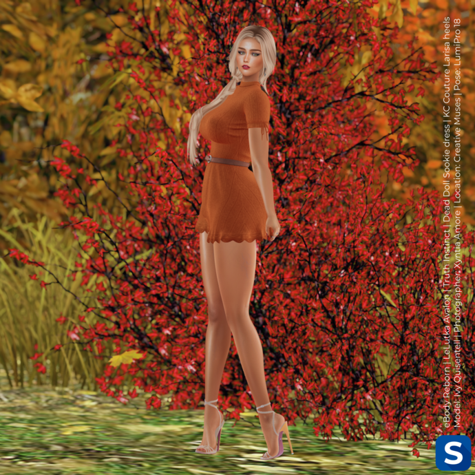 A full-length photo of Ivy Qissentell wearing an orange minidress while posed in front of some autumnal foliage. She has long blonde hair and is wearing high heels.