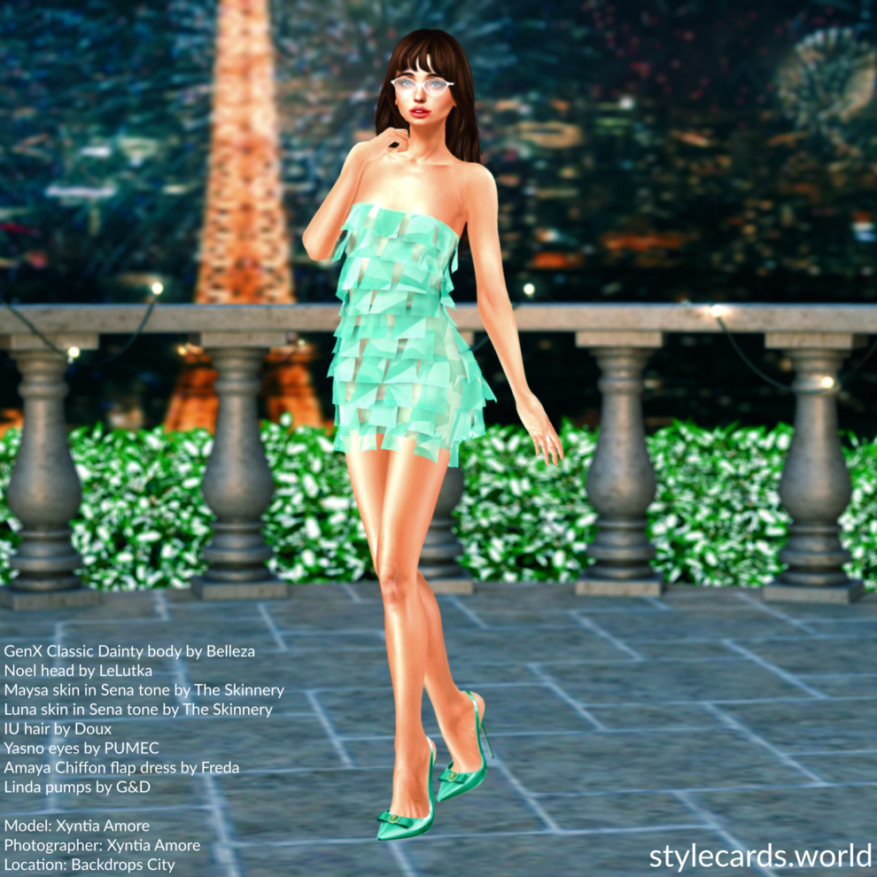 Xyntia Amore is standing on a patio in Paris at night as the Eiffel Tower shines brightly and fireworks illuminate the sky. She is wearing a teal Amaya Chiffon Flap dress from Freda and a pair of matching teal Linda pumps from G&D.
