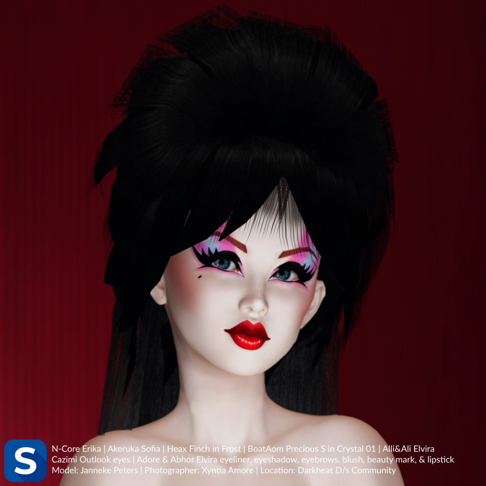 A headshot of Janneke Peters standing in front of a red wall. She has black hair in a beehive style hairdo with long straight hair down past she bare shoulders. She has arched eyebrows, pink and blue eyeshadow, black eyeliner and bright red lips, giving her the appearance of the character Elvira.