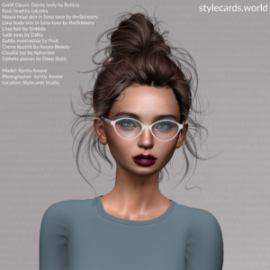 Stylecards Editor Xyntia Amore poses in front of a grey backdrop. She is wearing a slate blue long-sleeved t-shirt, has burgundy lipstick and pale blue eyeshadow. Her hair is done up in a bun, but is quite messy indicating her hard work. She is wearing a pair of white-framed eyeglasses.