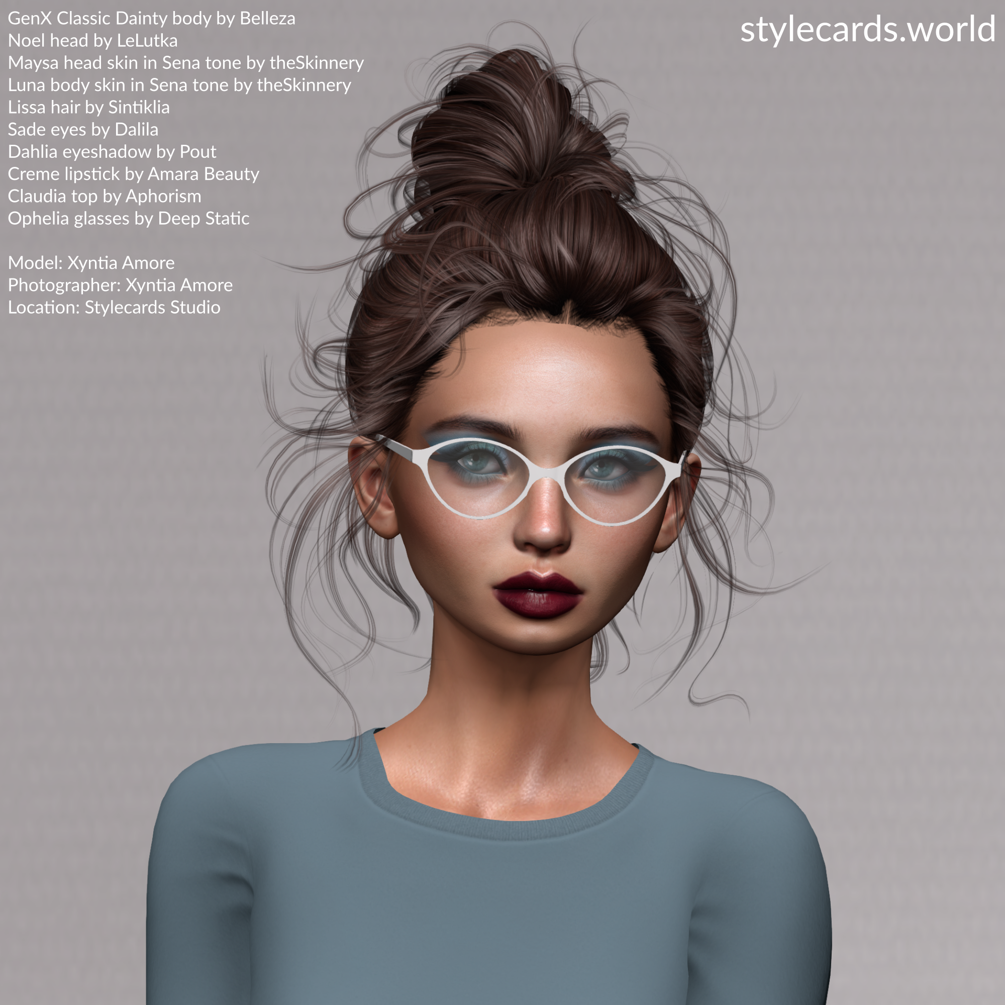 Stylecards Editor Xyntia Amore poses in front of a grey backdrop. She is wearing a slate blue long-sleeved t-shirt, has burgundy lipstick and pale blue eyeshadow. Her hair is done up in a bun, but is quite messy indicating her hard work. She is wearing a pair of white-framed eyeglasses.