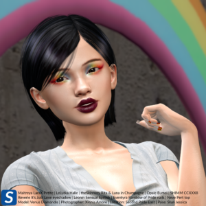 Venus Diamandis is standing in front of a rainbow prop while wearing Pride eyeshadow and fingernails. She has short black hair and is wearing a pale blue blouse.