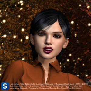 A headshot of Venus Diamandis has her standing in front of some trees bearing leaves in autumnal colors, and which coordinate well with her dark orange polo shirt. She is wearing dark red lipstick, black eyeliner, and subtly smokey eyeshadow. Her hair is short and black.