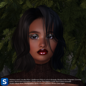 A headshot of model Kalyani Bose as she stands in front of a pine tree at a ski lodge. She has shoulder length wavy black hair and is wearing a brown sweater. Her lips are bright red and her eyeshadow is a mix of silver and green with Christmas tree decorations adorning her right upper lid.