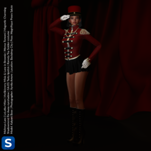 Kalyani Bose stands and salutes in front of some red curtains. She is wearing black high heel boots that reach above her calves, a black miniskirt, a red military-style uniform jacket trimmed with black and gold details, with a matching red, black, and gold shako atop her head.