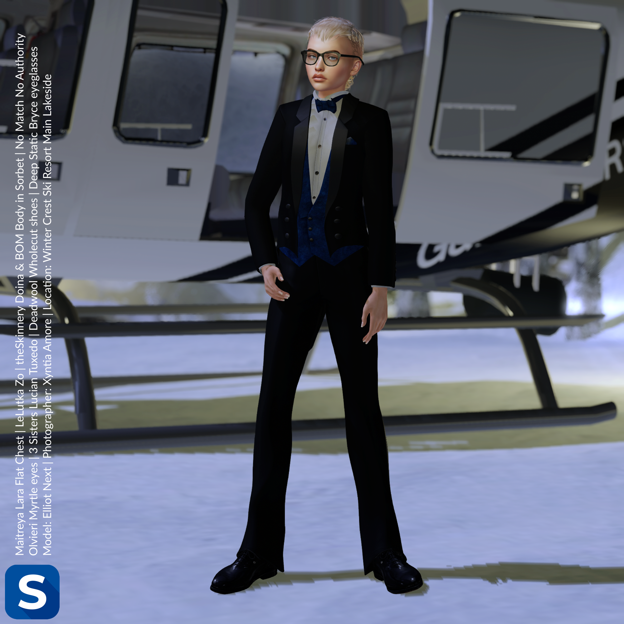 Model Elliot Next is wearing a black tuxedo with blue accents while standing next to a helicopter at a ski resort. His feet a shod in black leather dress shoes and he has black framed eyeglasses on his face. His hair is short and blond.