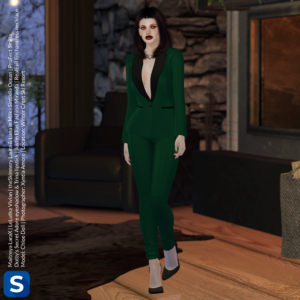 A photo of model Chloe Doll wearing a green tuxedo with black accents, and accessorized with black high heel shoes and a black metal necklace in a form inspired by antlers. Behind her is a fireplace and a wall made of stone. Through a nearby window we can see snow falling outside.
