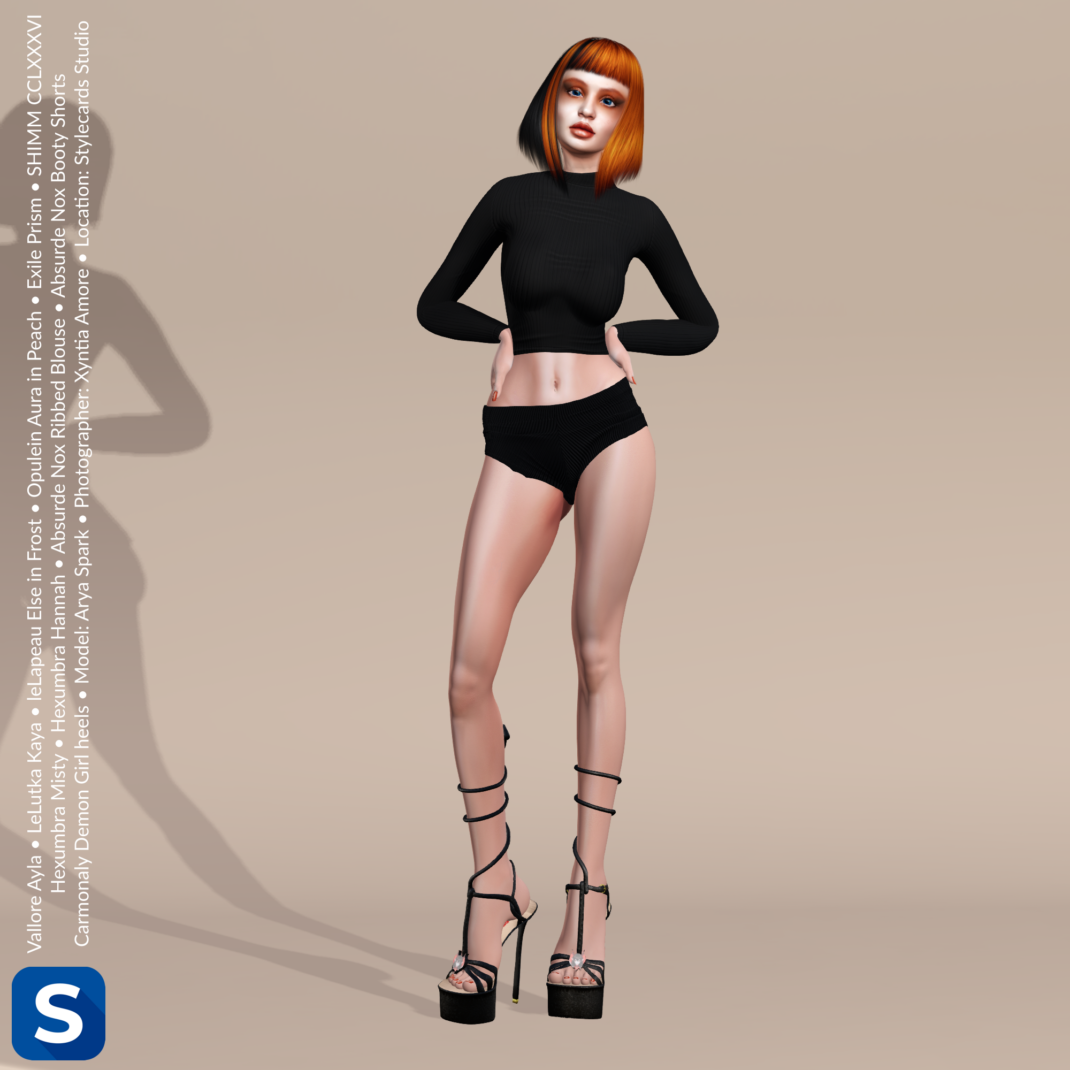Threadsday, August 13, 2026 A full shot of Arya Spark standing in front of a beige backdrop. She is wearing a black long-sleeved cropped top, black booty shorts and black strappy heels. Her hair is short and makes her look like a redhead except for the black streaks. Her eyeshadow and lipstick are orange.