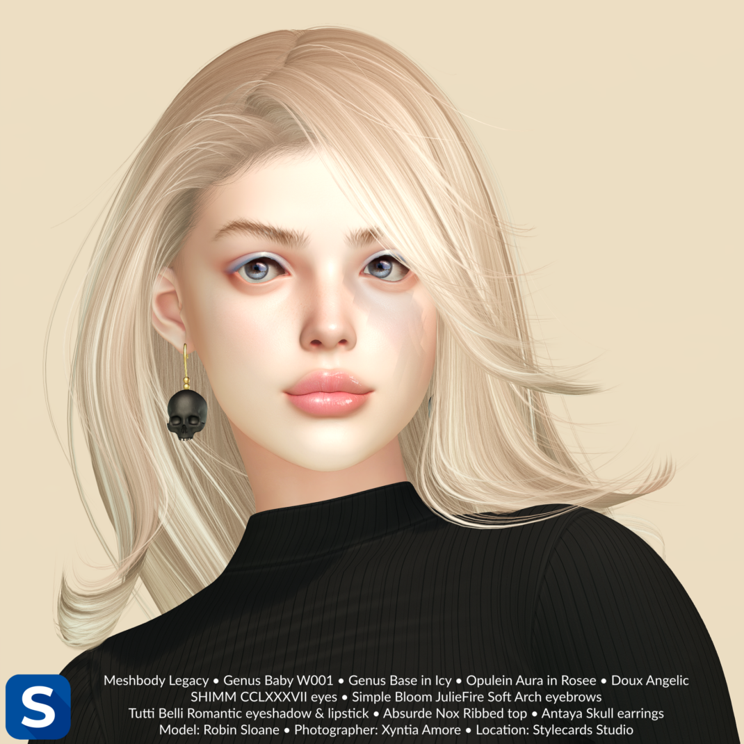 A headshot of a woman with long blonde hair standing in front of a pale yellow backdrop. She has pale blue eyeshadow, pale pink lipstick, and skull-shaped earrings in black to match the color of her sweater.