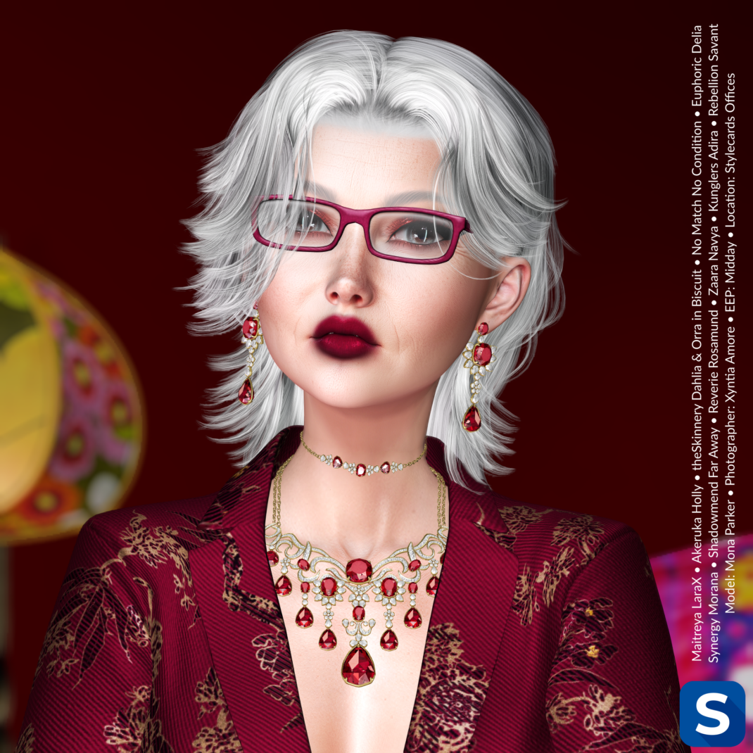 Makeup Monday, September 21, 2026 A headshot of a woman with shoulder-length silver hair. She is wearing a suit jacket that is red brocade with gold-colored floral motifs, A gold necklace with several red gemstones is draped from her neck and matching earrings hang from her ears. Behind her is a red wall and some multi-colored items of furniture.