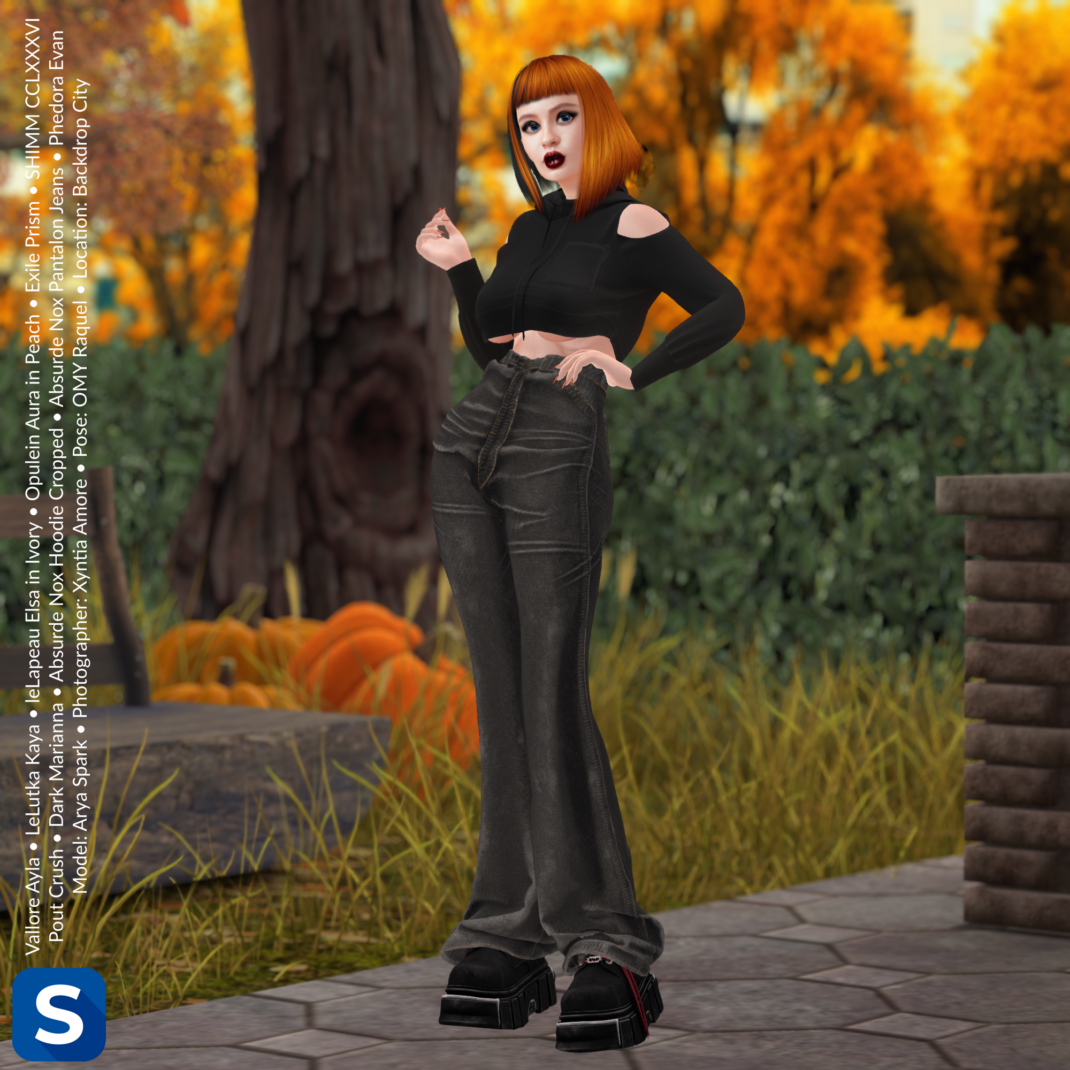 Threadsday, September 17, 2026 A female model wearing a black cropped hoodie and black denim jeans is standing in a park with trees in the background that have leaves that are bright yellow and orange. Her short hair is bright orange with a black streak and she has black boots on her feet.